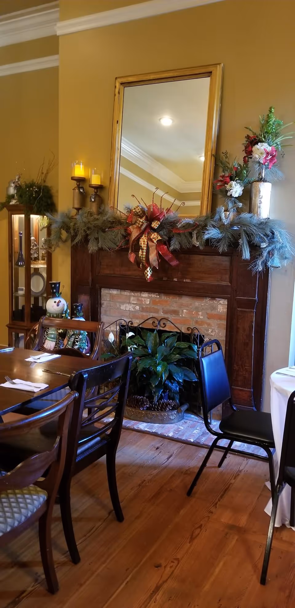 A cozy dining area with wooden tables and chairs, featuring a decorated fireplace mantel adorned with greenery, candles, and a festive bow. A large mirror hangs above the mantel, reflecting ceiling lights. There is a plant inside the fireplace and a snowman decoration on a side table.