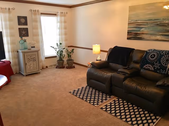 Cozy living room with a leather reclining sofa, small area rugs, side table with a lamp, potted plants, and wall art near a window.