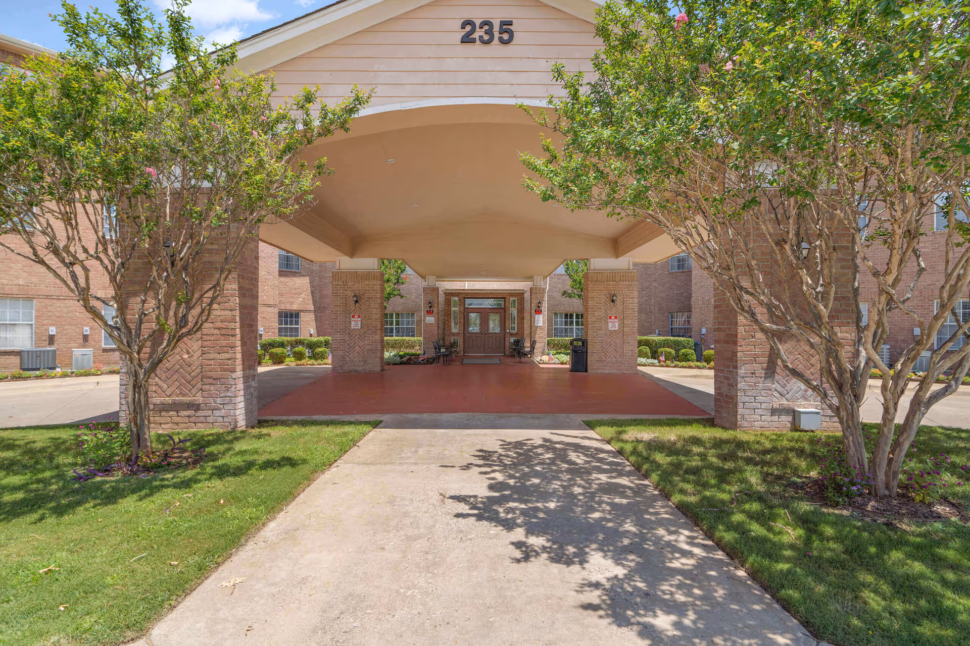 Entrance to a senior living facility with a covered driveway supported by brick pillars, flanked by green trees and shrubs, with the number 235 displayed above the entrance.