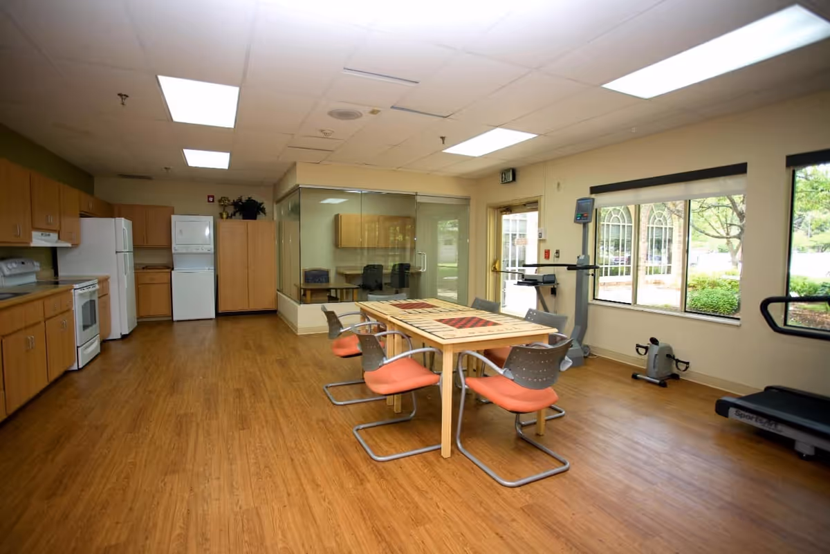 Spacious communal room with a kitchenette along the left wall, a central table surrounded by orange chairs, and exercise equipment by large windows.