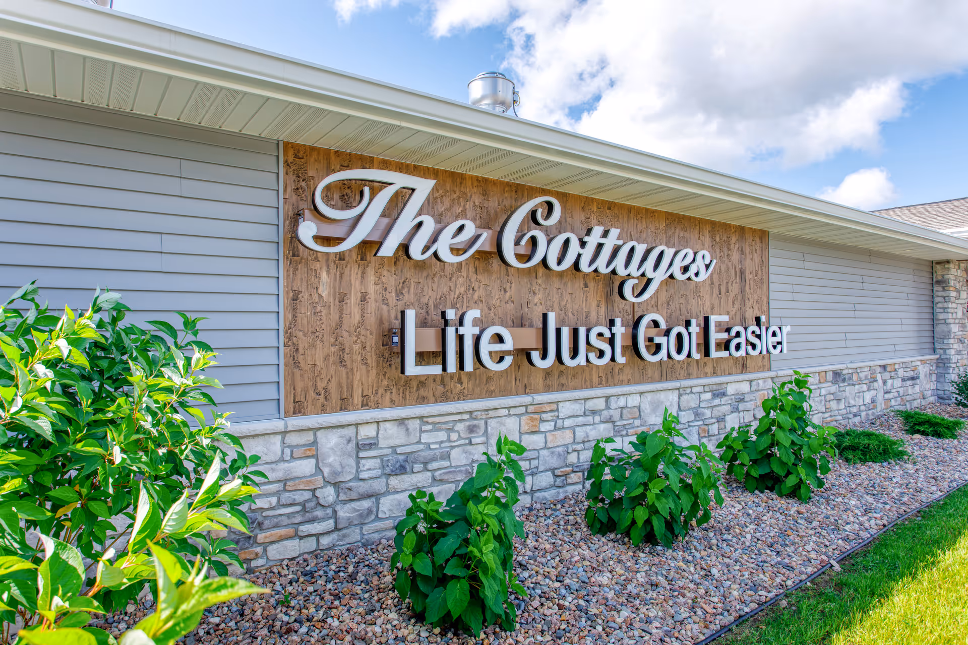 Exterior view of a building wall with a large sign that reads 'The Cottages Life Just Got Easier' mounted on a wooden panel above a stone facade, with green plants and a blue sky with clouds in the background.
