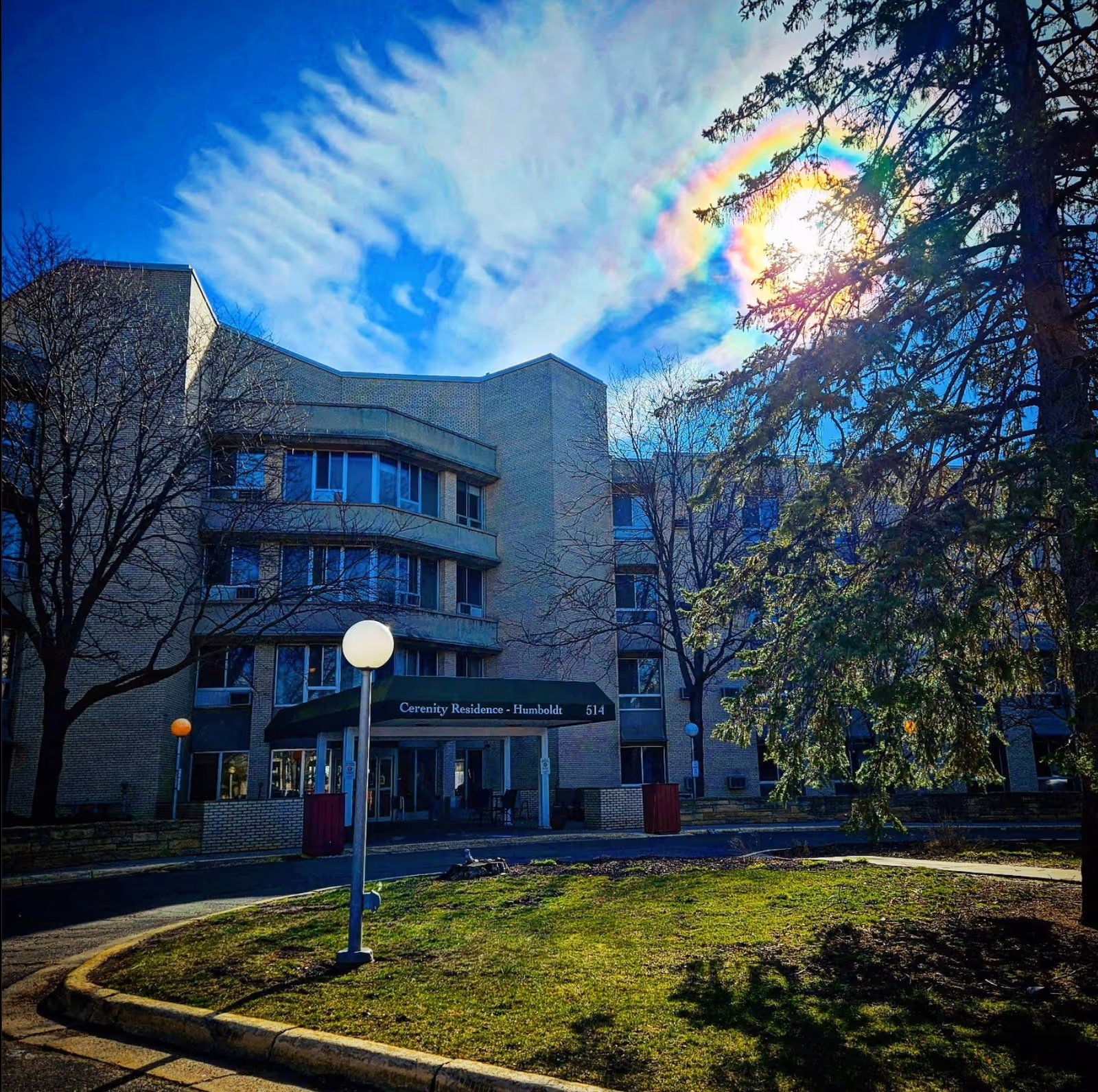 Multi-story beige senior living building with curved façade and entrance canopy under a rainbow halo.