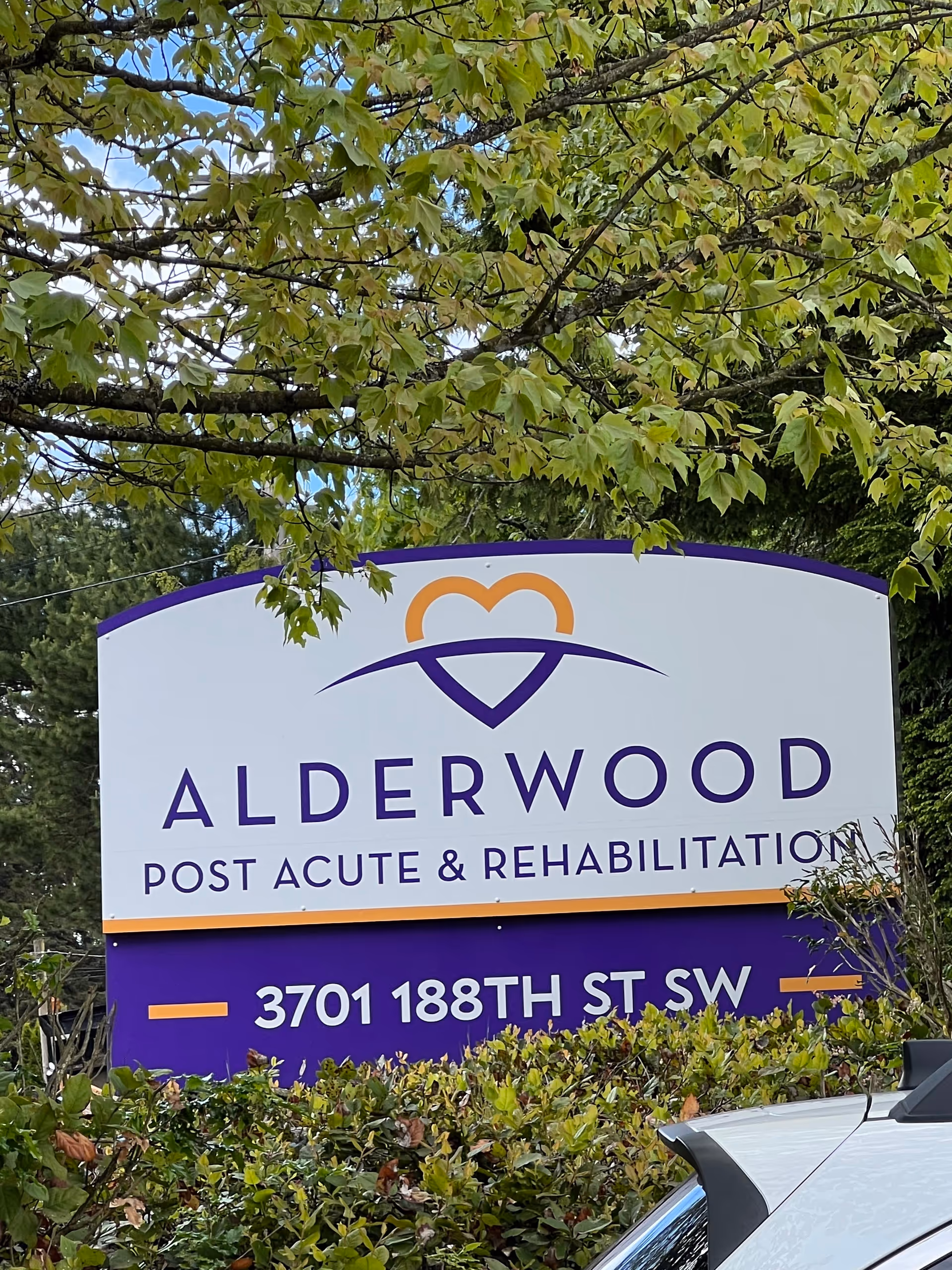 Outdoor sign reading Alderwood Post Acute & Rehabilitation with address, surrounded by shrubs.