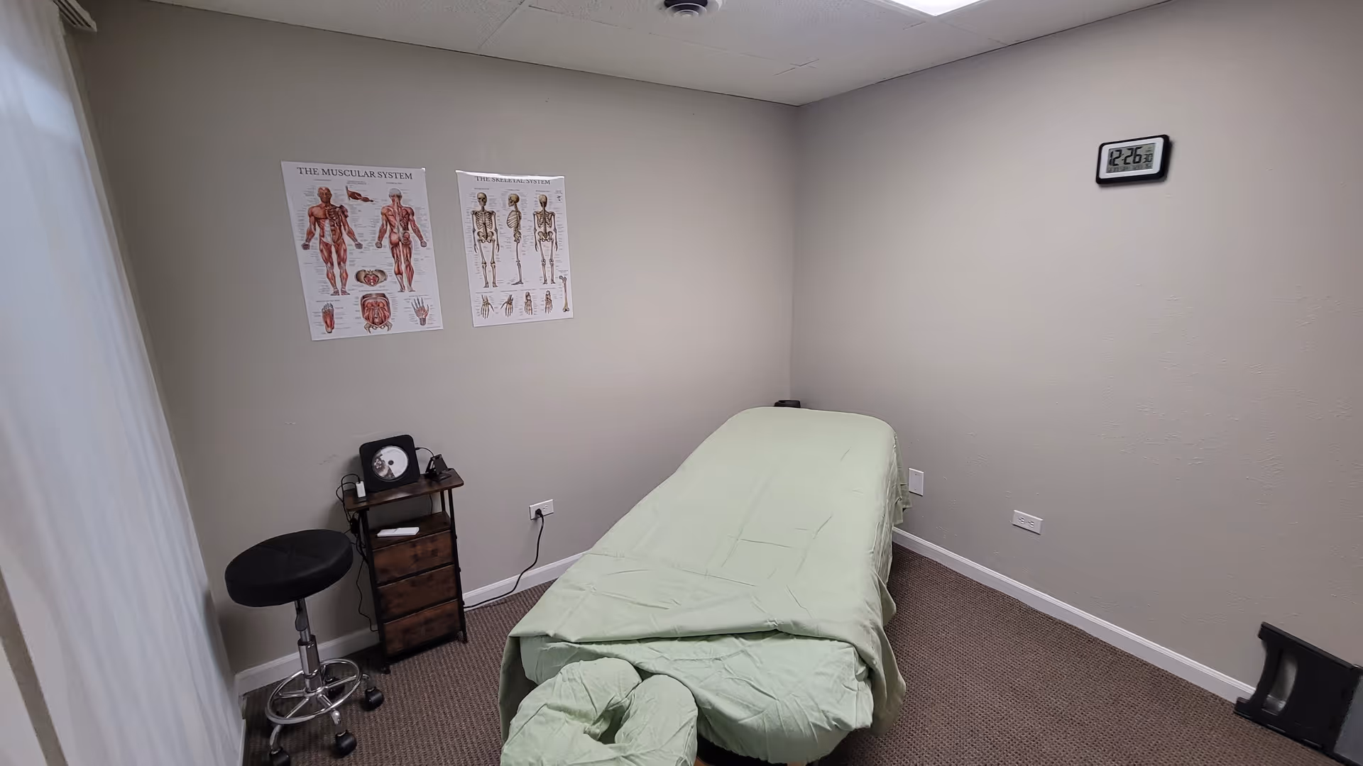 A massage therapy room with a table, posters, a stool, and a wall clock.