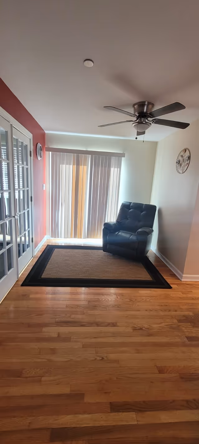 A small living area with a black leather recliner on a rug before vertical blinds, hardwood floors, a ceiling fan, and glass French doors.