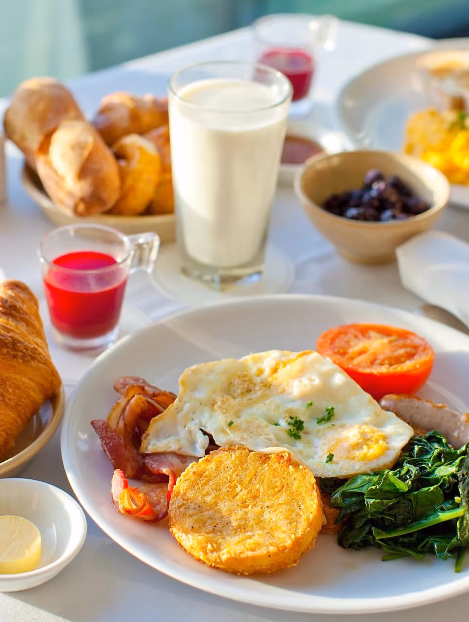 Close-up breakfast spread on a dining table with eggs, croissant, milk, juice, and sides.