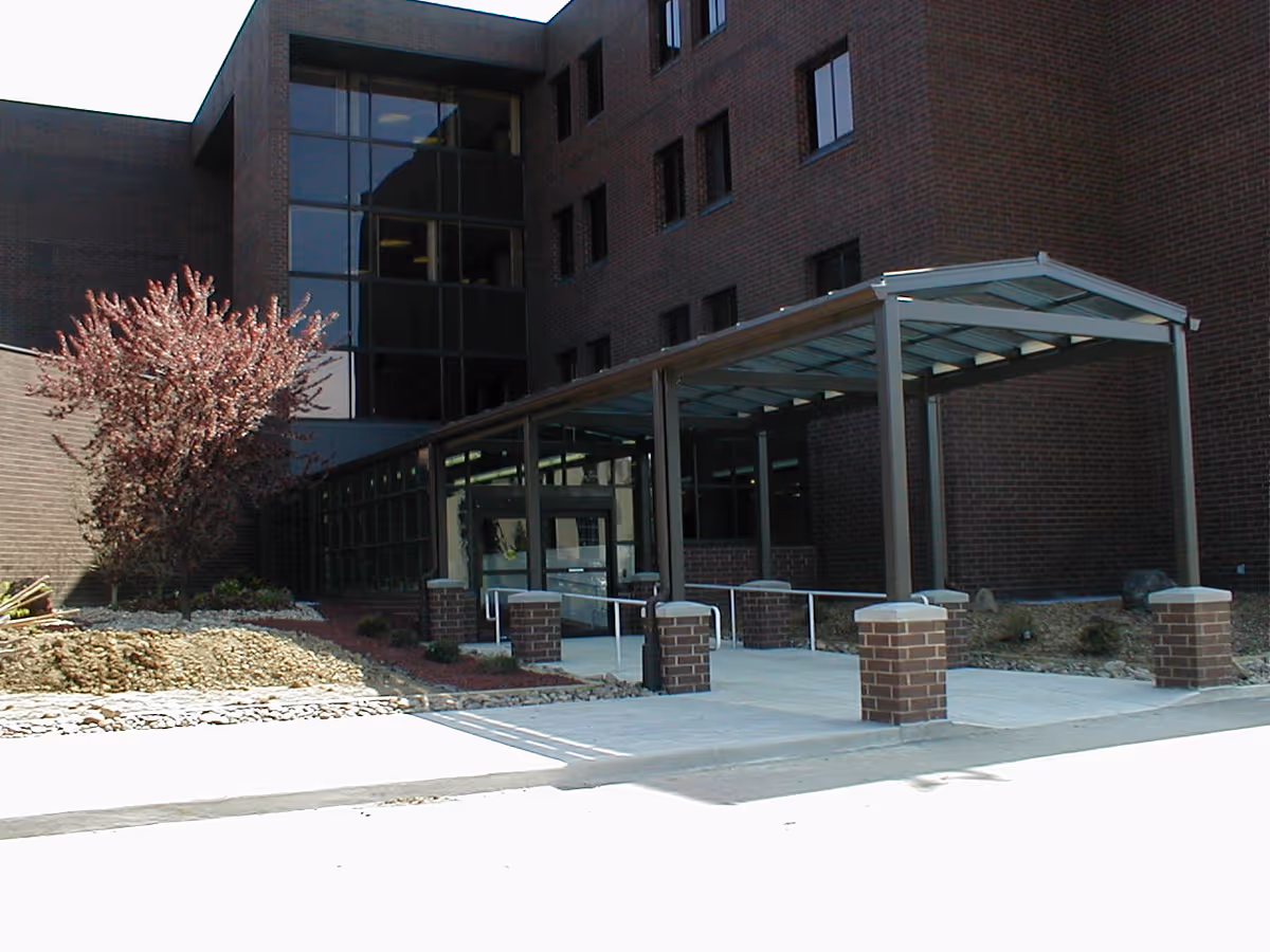 Exterior view of Westmoreland Manor showing a brick building with multiple windows and a covered walkway supported by brick pillars leading to the entrance. There is a small landscaped area with a tree and rocks beside the walkway.