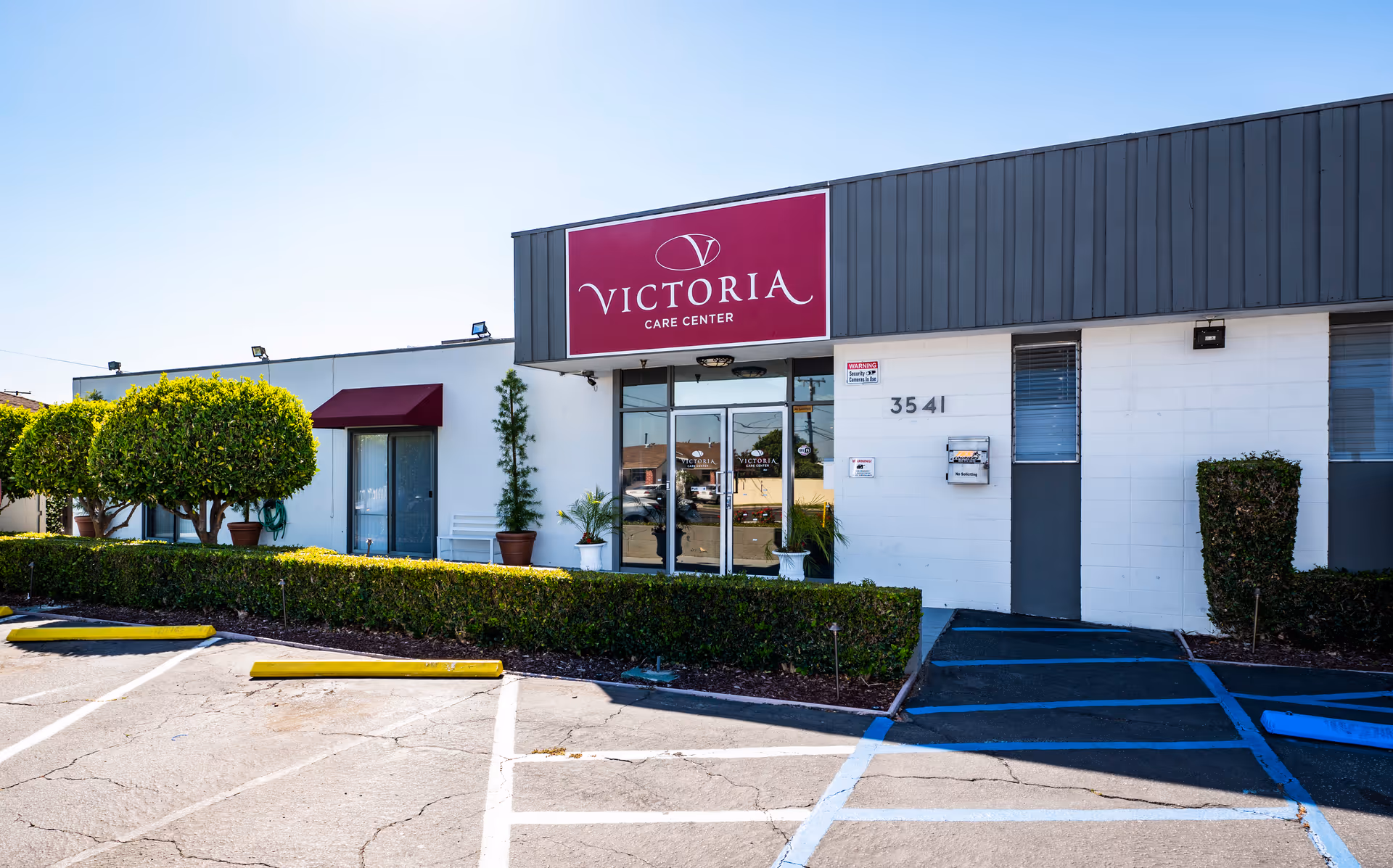 Exterior view of Victoria Care Center building with a large maroon sign above the entrance. The building is white with a gray upper facade, surrounded by trimmed bushes and small trees. There is a parking lot with marked spaces and a handicap accessible ramp leading to the entrance.