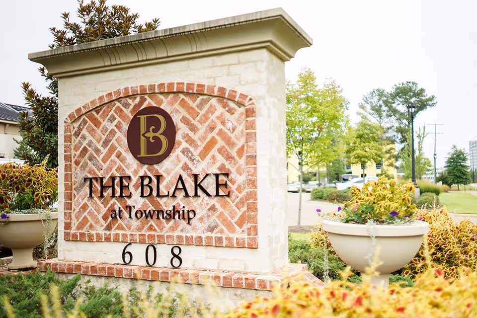 A brick and stone sign for The Blake at Township with the number 608 at the bottom, surrounded by greenery and flowers in planters, with trees and a street visible in the background.