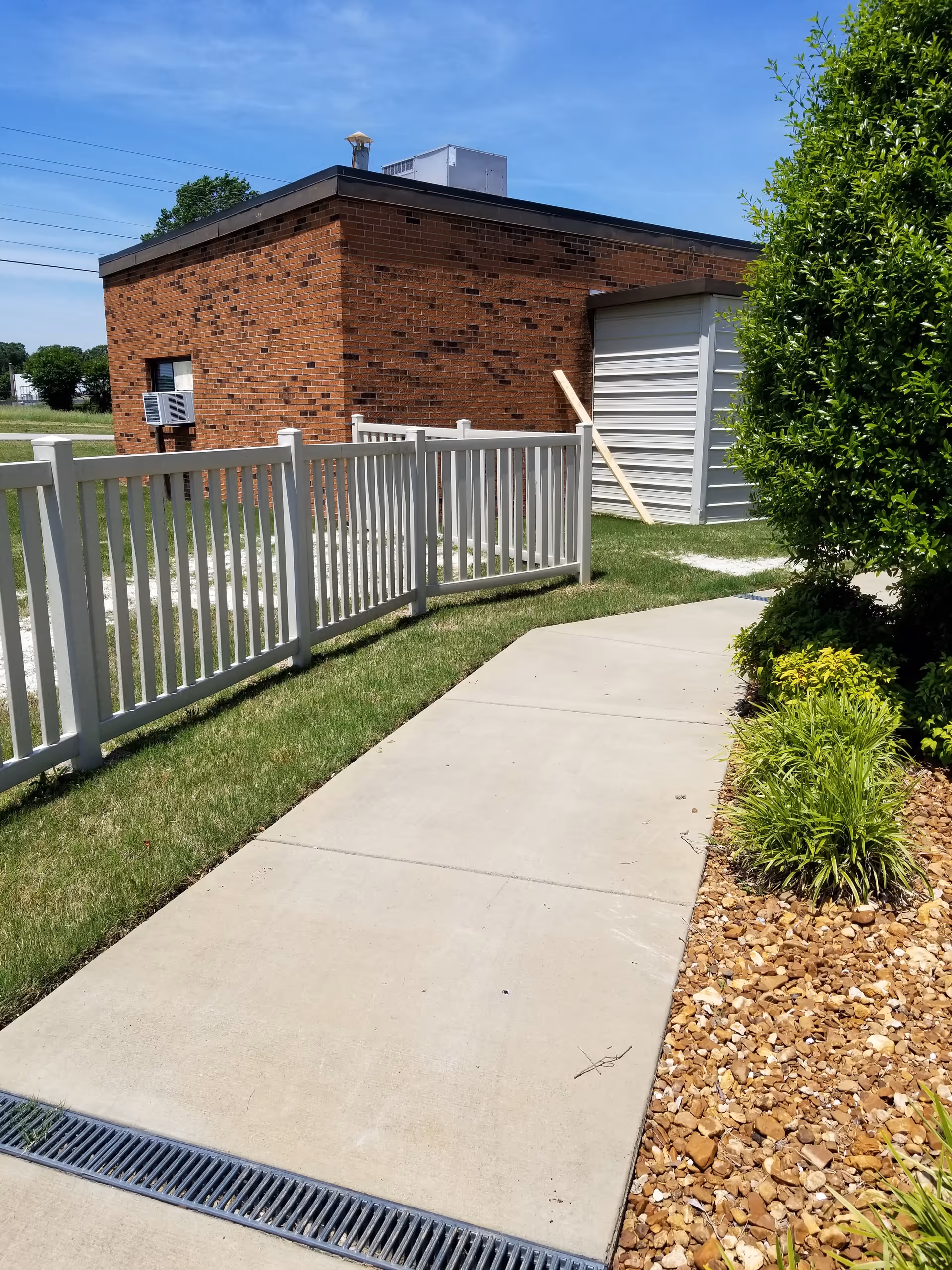 Exterior view of a brick facility with a white fence, sidewalk, and landscaped area.