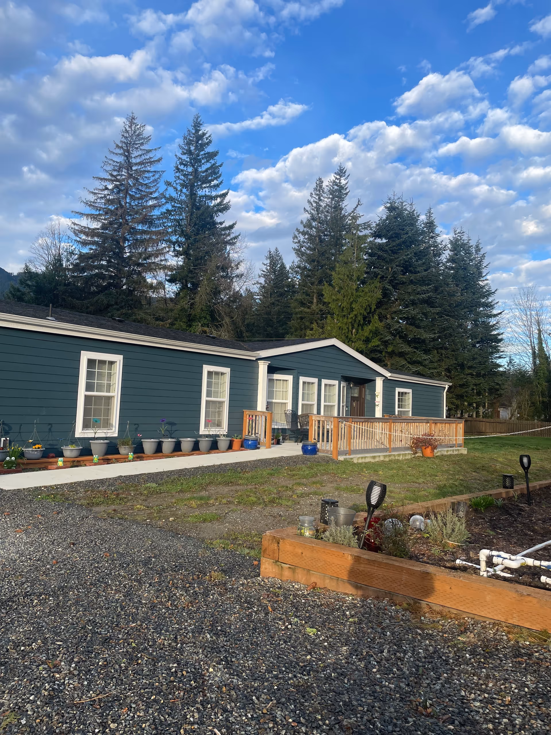 Blue single-story care facility with a wooden deck, planters, and a gravel yard outdoors.