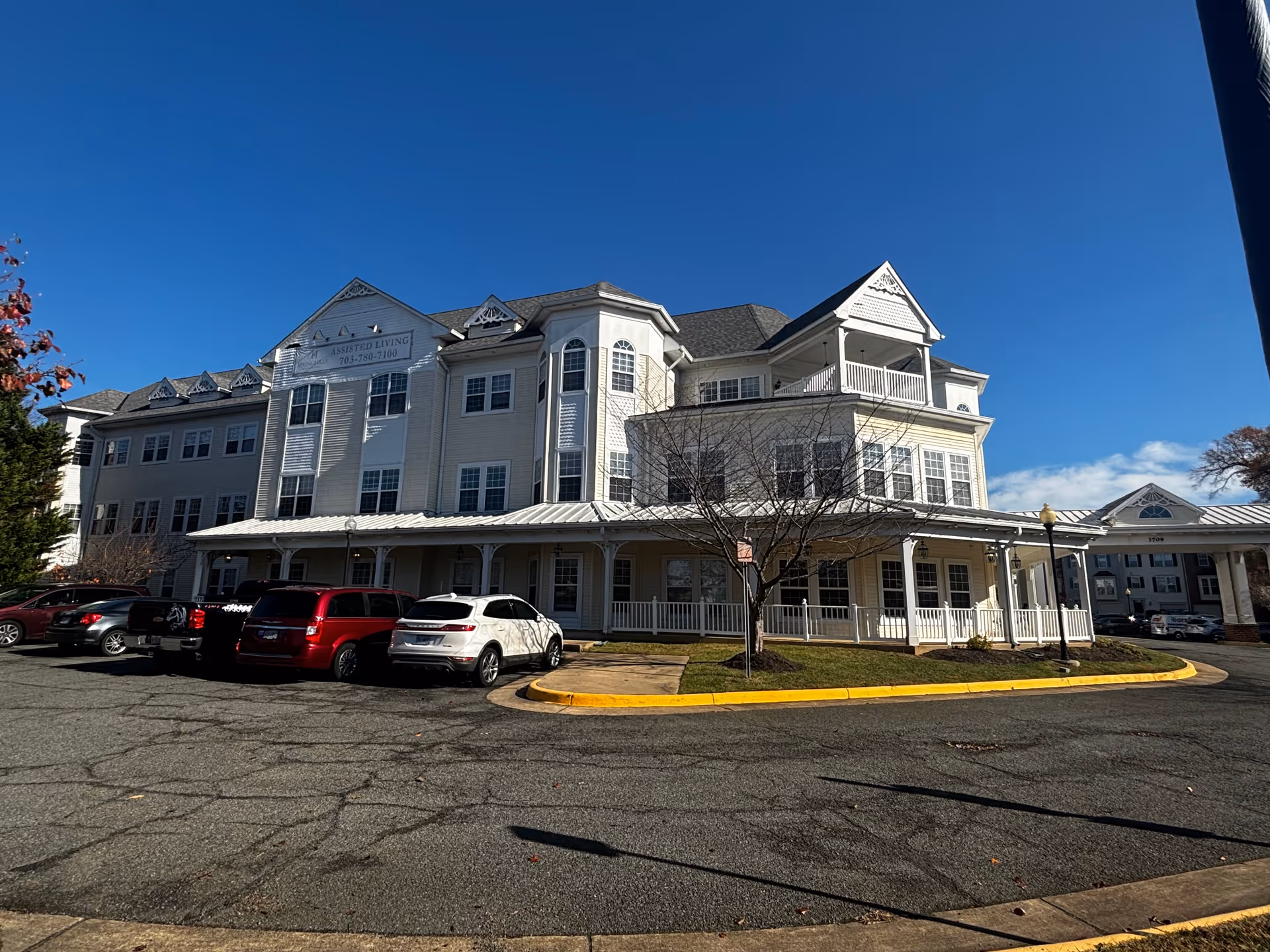 Front exterior of a multi-story senior living building with cars parked in front under a clear blue sky.