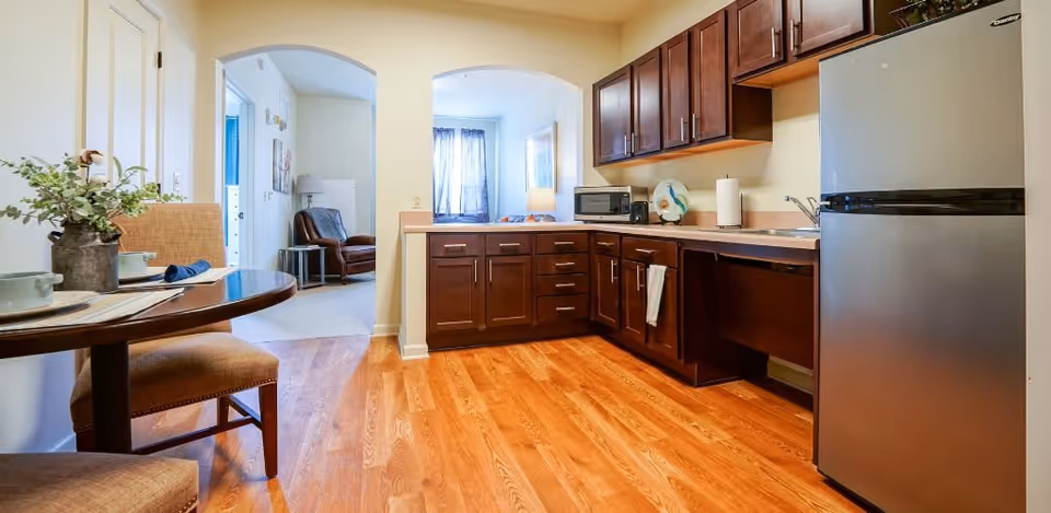 A bright kitchen area with wooden cabinets, a stainless steel refrigerator, a microwave, and a small dining table with two chairs. The floor is wooden, and through two archways, a living area with a leather chair and a window with curtains is visible.