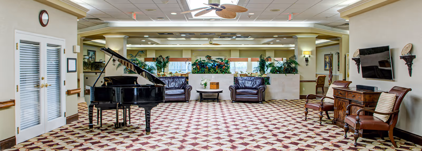 A spacious and well-lit common area in a senior living facility featuring a black grand piano in the center, two dark leather sofas with a round coffee table between them, patterned carpet flooring, several potted plants, a wall-mounted flat-screen TV with two chairs and a wooden cabinet beneath it, and ceiling fans above.
