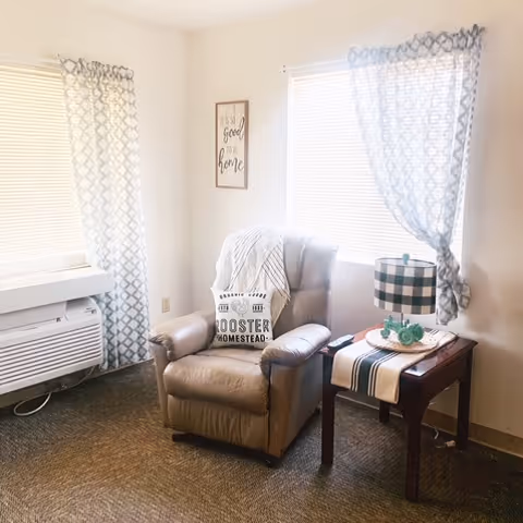 Cozy senior living room corner with armchair, side table, lamp, and patterned curtains.