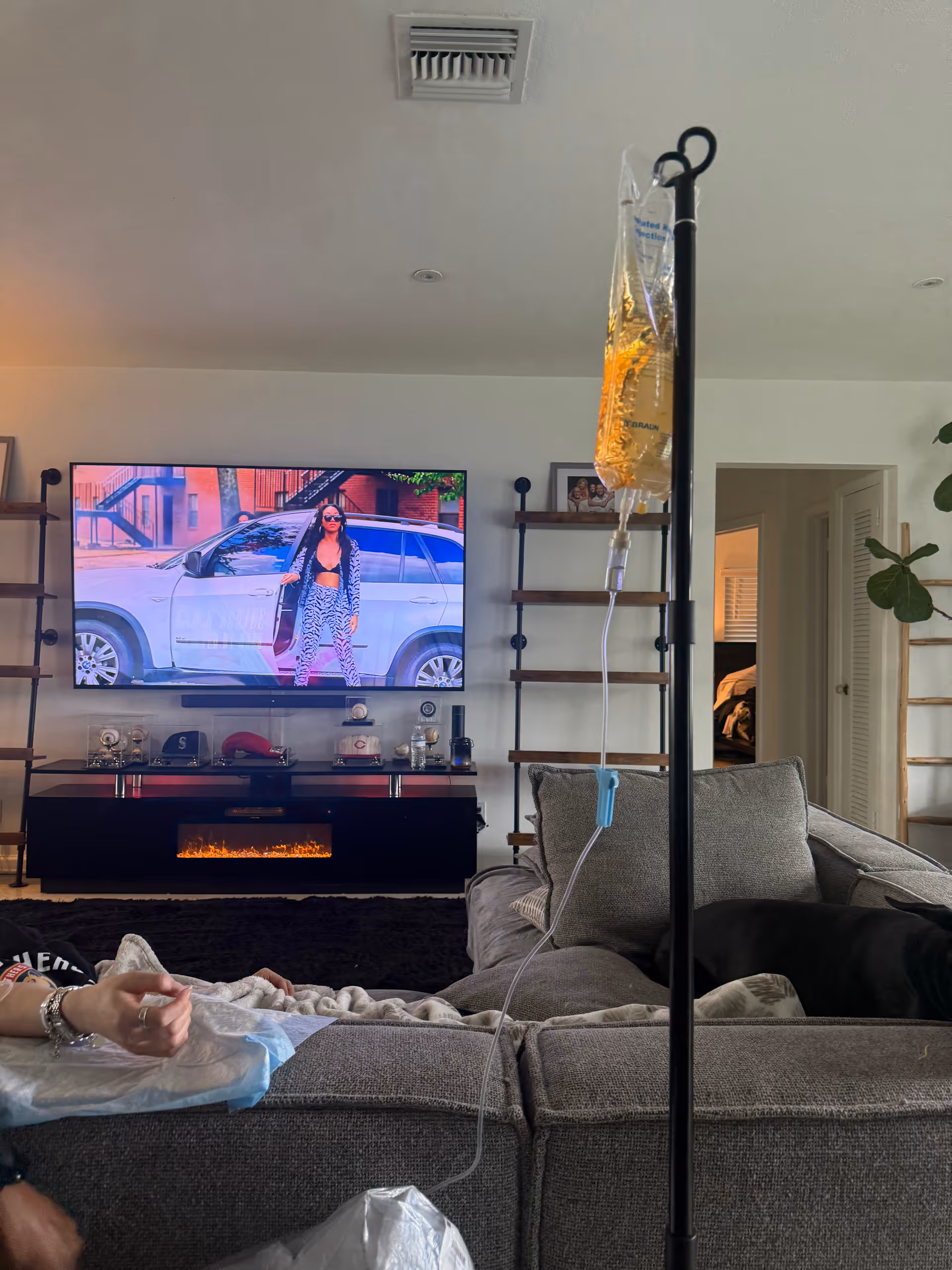 A living room with a gray sectional, mounted TV, and an IV drip stand visible.