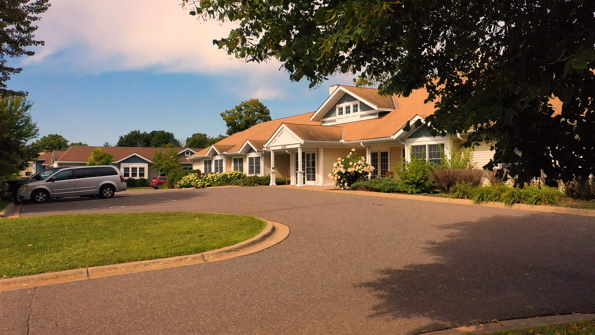 Exterior view of a single-story senior living facility building with a tan roof and beige siding, surrounded by greenery and trees, with a paved driveway and parked vehicles nearby under a partly cloudy sky.