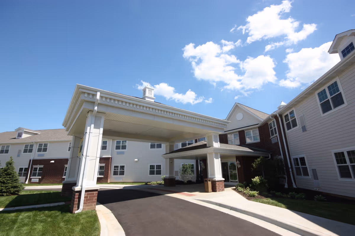 Exterior view of a senior living facility building with a covered entrance driveway, white siding, brick accents, multiple windows, and a clear blue sky with some clouds.
