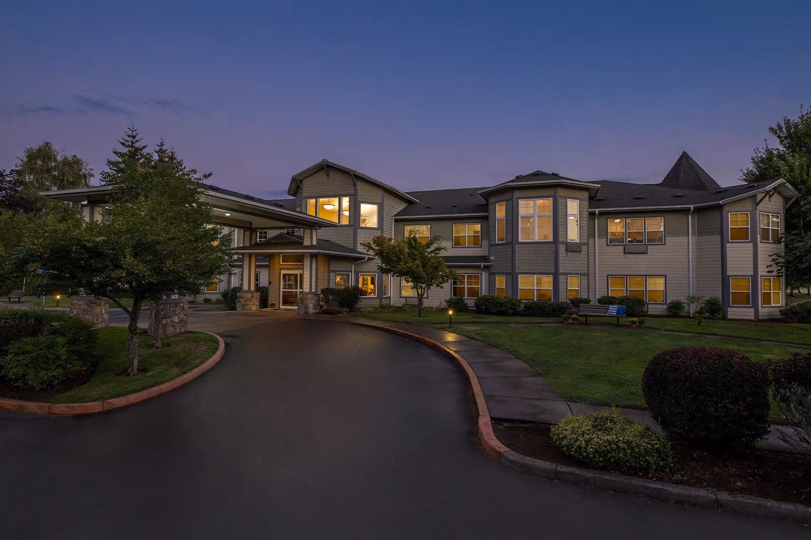 Exterior view of a senior living building with a curved driveway and lit windows at dusk.