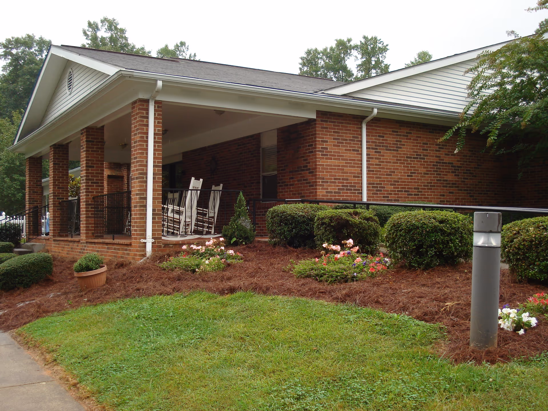 Brick building front with a covered porch featuring rocking chairs, landscaped bushes and flowerbeds.