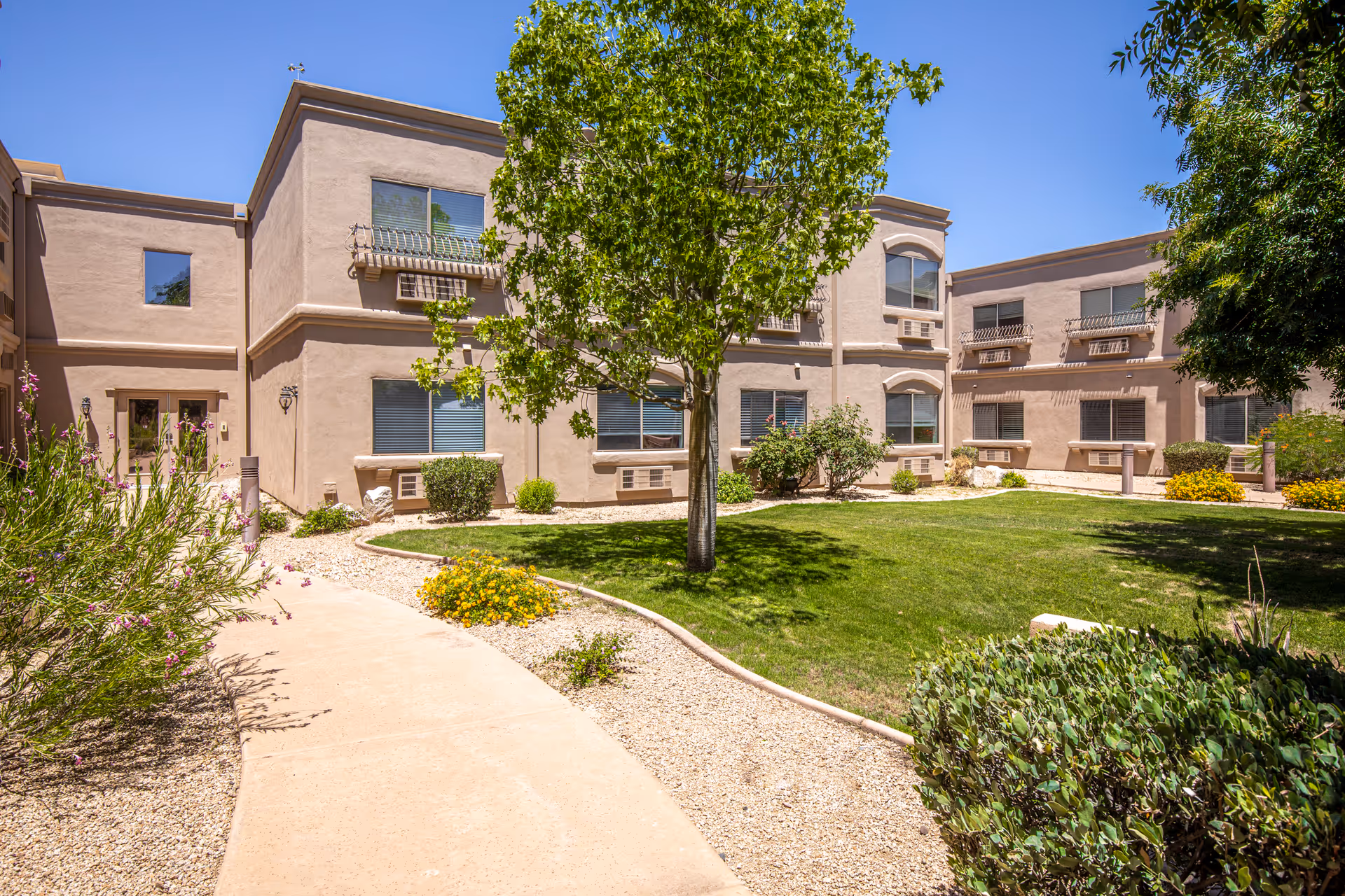 Courtyard with a paved walkway, grassy lawn and trees surrounded by a two-story beige residential building.