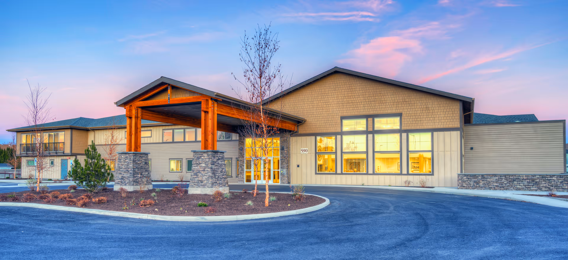 Exterior view of Juniper Springs Senior Living facility at dusk, showing a modern building with large windows, a covered entrance supported by wooden beams and stone pillars, surrounded by a paved driveway and landscaped areas with small trees and shrubs.
