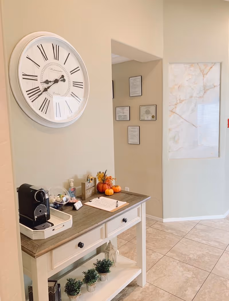 Interior entryway with a console table holding a coffee machine, small potted plants and fall decor, a large wall clock above, and framed certificates on the wall.