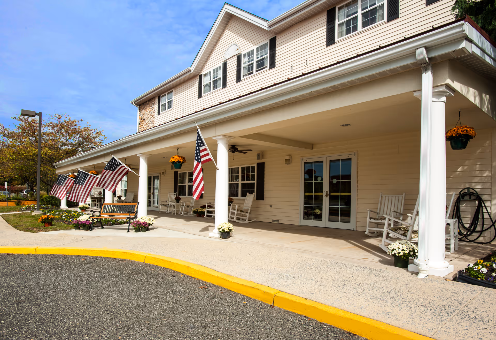Front exterior view of a senior living facility with a covered porch featuring white columns, rocking chairs, hanging flower pots, American flags, and a bench surrounded by flower beds under a clear blue sky.