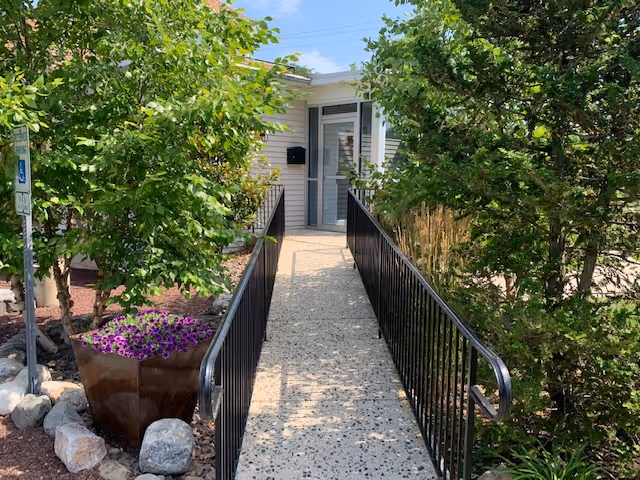 A concrete ramp with black railings leads to a building entrance surrounded by trees and planters.