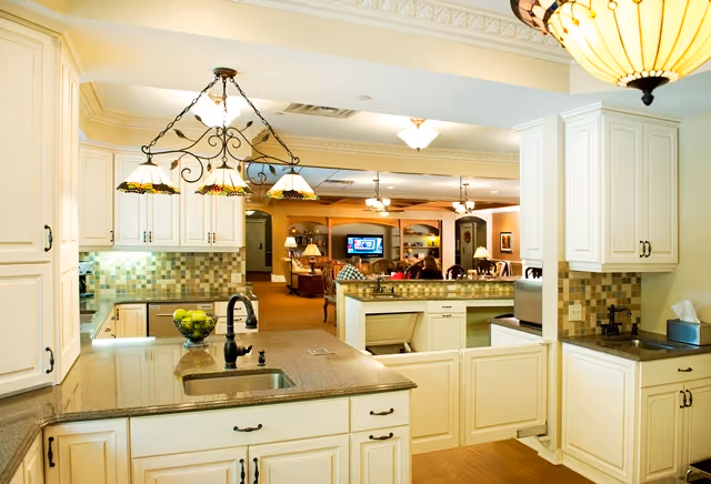 Bright kitchen with white cabinets, granite island, tiled backsplash, and open lounge in background.