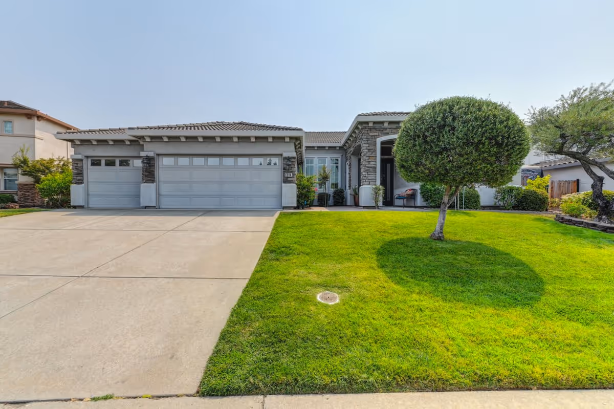 Front exterior of a single-story suburban house with a three-car garage, concrete driveway and a well-manicured lawn featuring a round topiary tree.