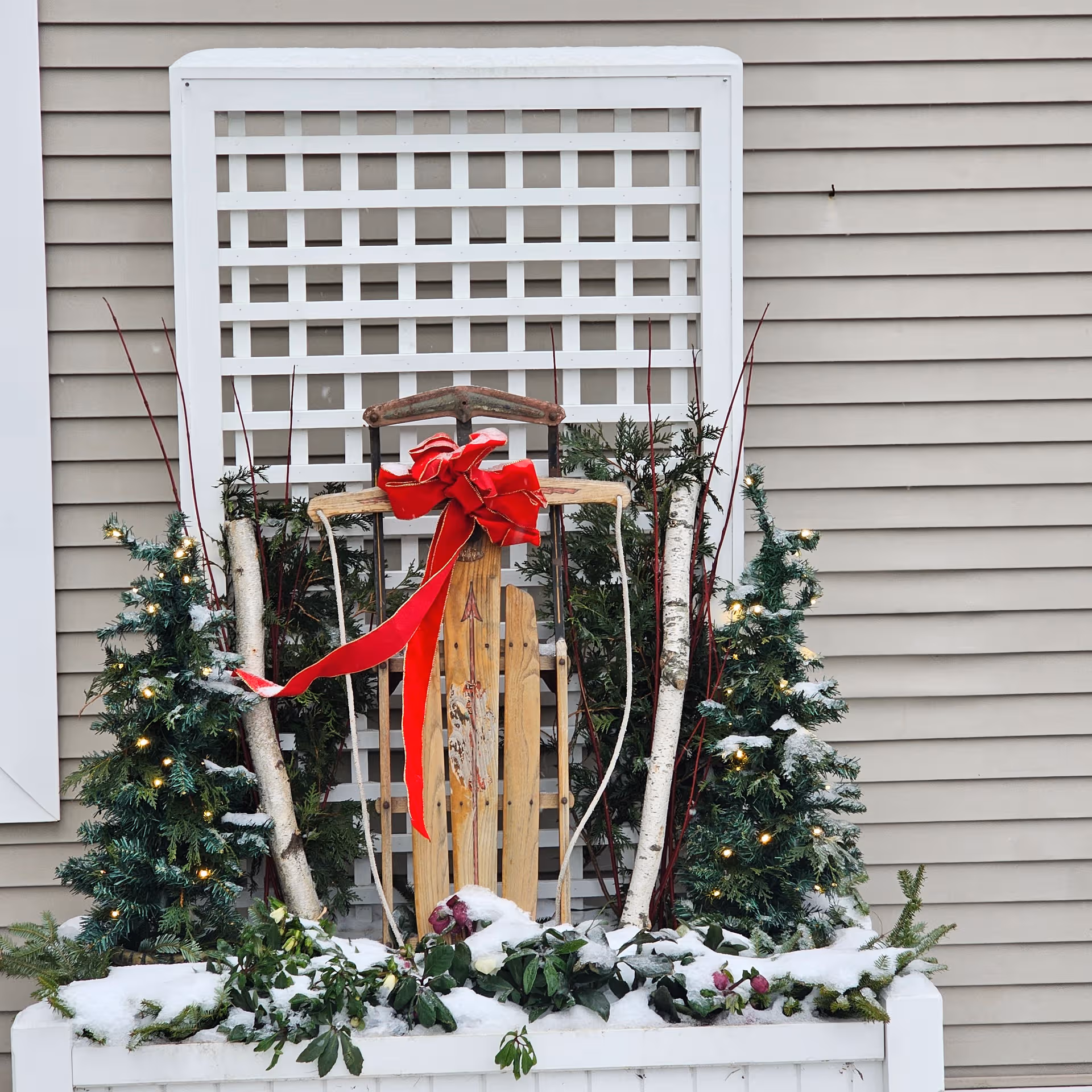 Winter holiday display with a wooden sled, red bow, greenery, and snow in front of siding.
