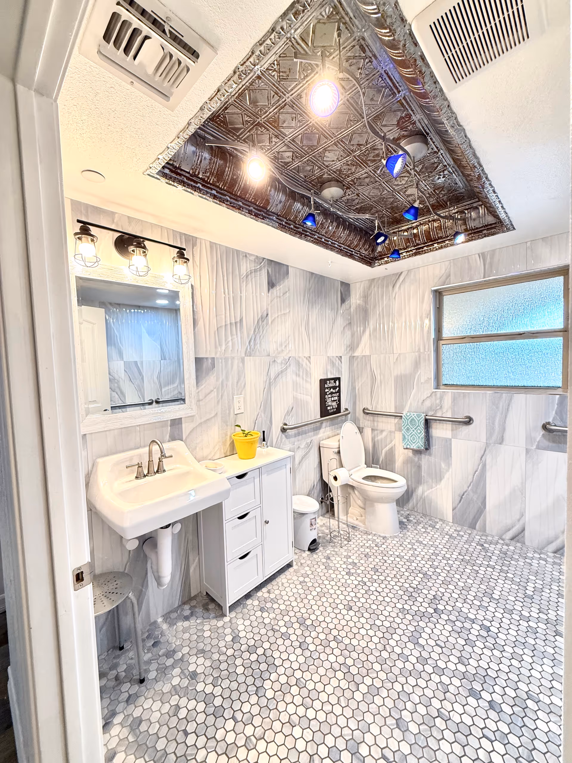 Modern tiled bathroom with sink, toilet, vanity, grab bars, and blue-lit ceiling lights.