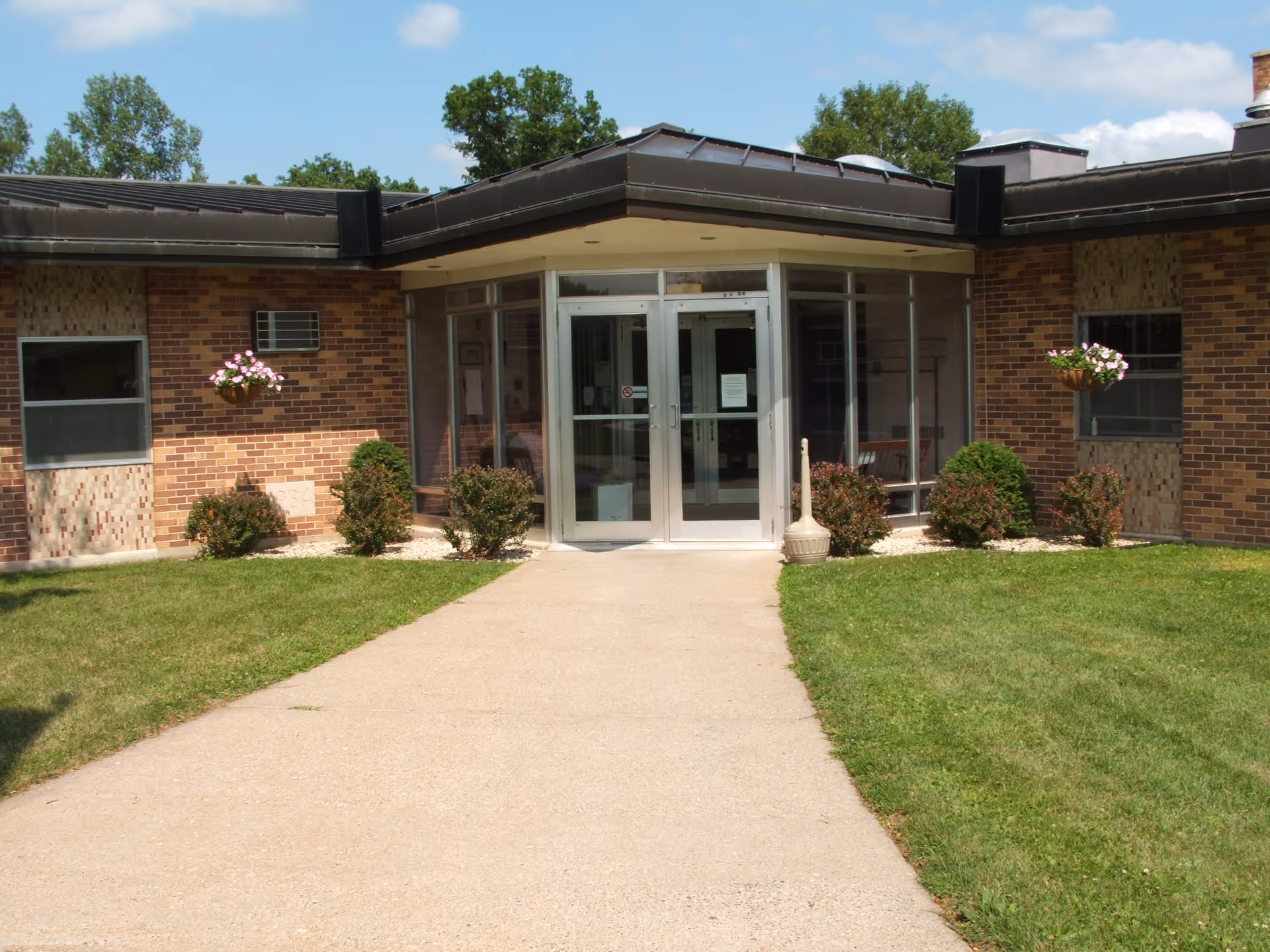 Entrance of a brick building with glass double doors, surrounded by small bushes and hanging flower pots on either side. A concrete pathway leads up to the entrance, with green grass on both sides and trees in the background under a partly cloudy sky.