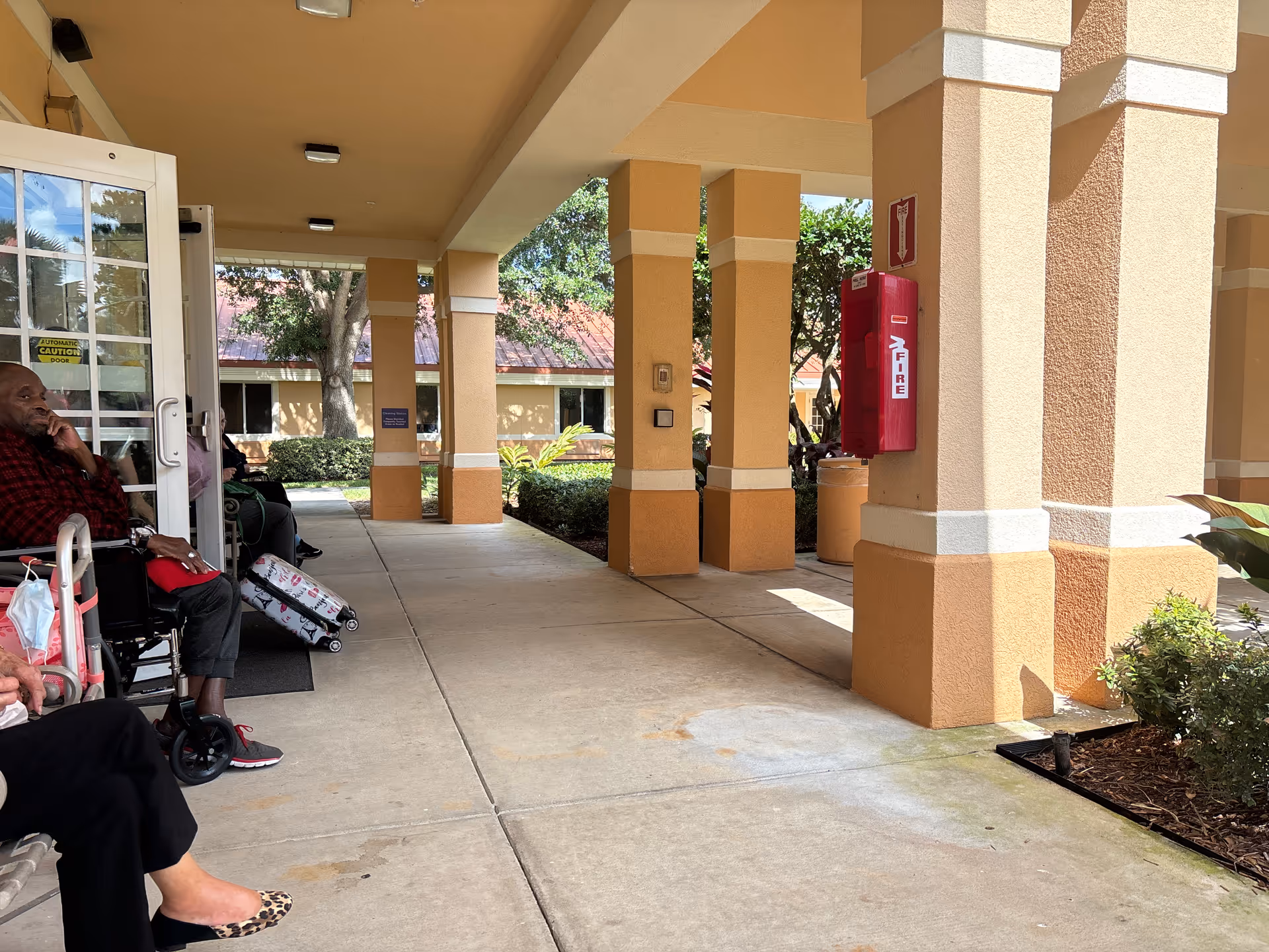 Residents sit in wheelchairs along a covered entrance walkway at a senior care facility.