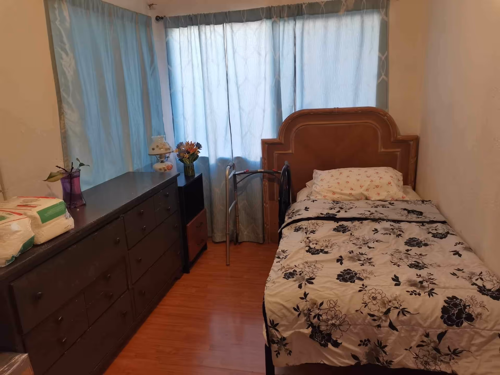 Small bedroom with a single bed, floral quilt, dresser, and blue curtains.