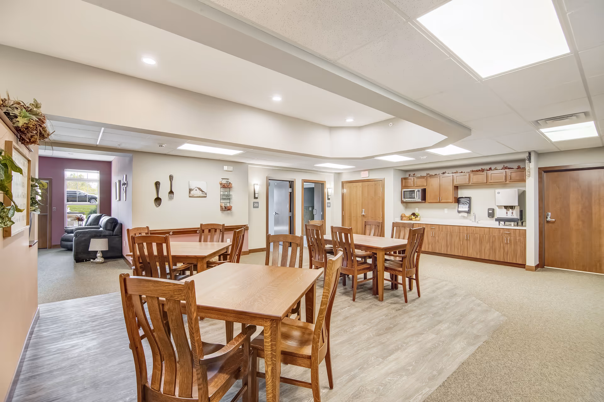 Bright communal dining area with wooden tables and chairs and a kitchenette in a senior living facility.