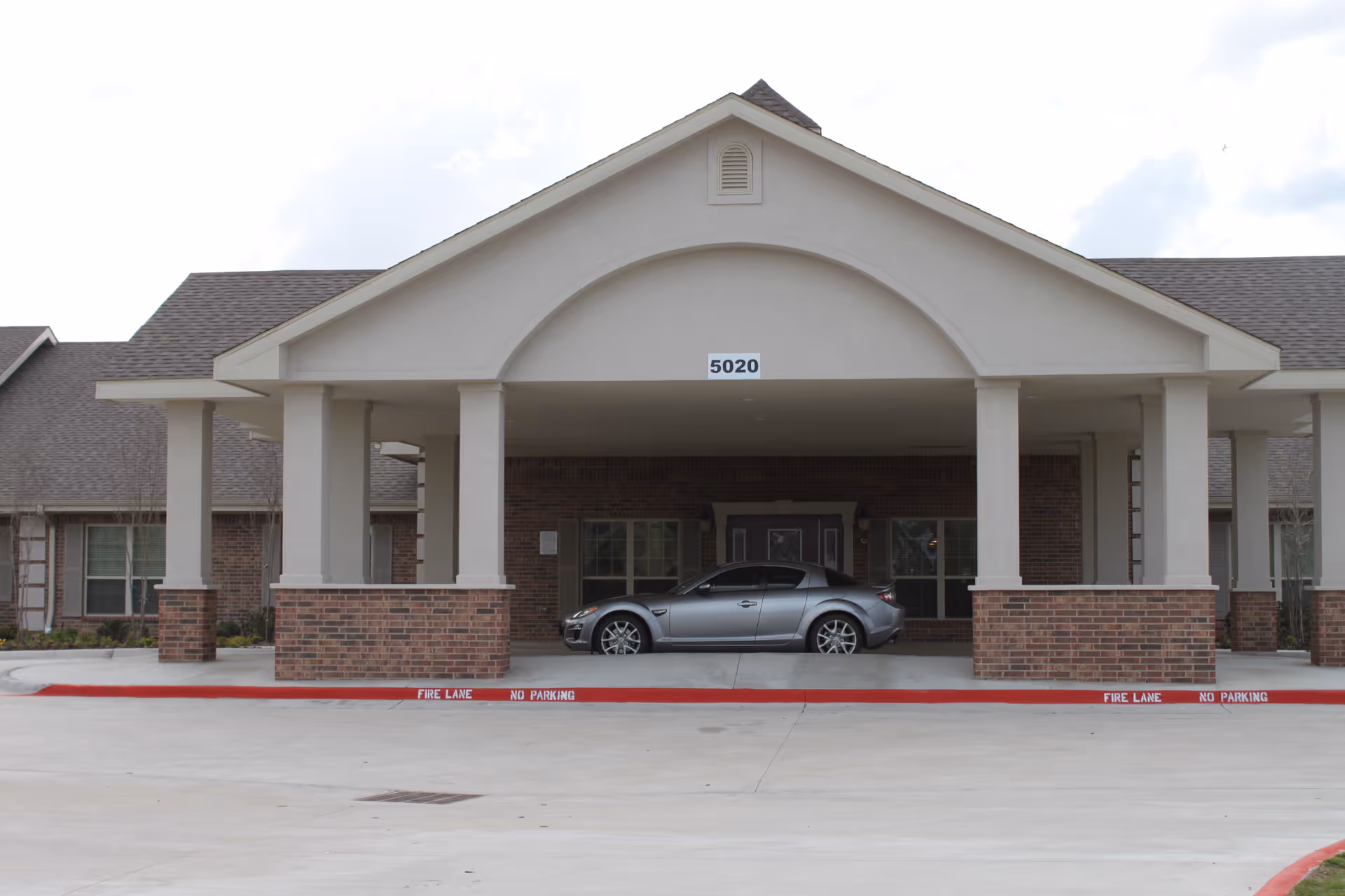 Front exterior view of a building with a covered entrance supported by columns. A silver car is parked under the covered area. The building has brick and beige walls with a sign displaying the number 5020 above the entrance.