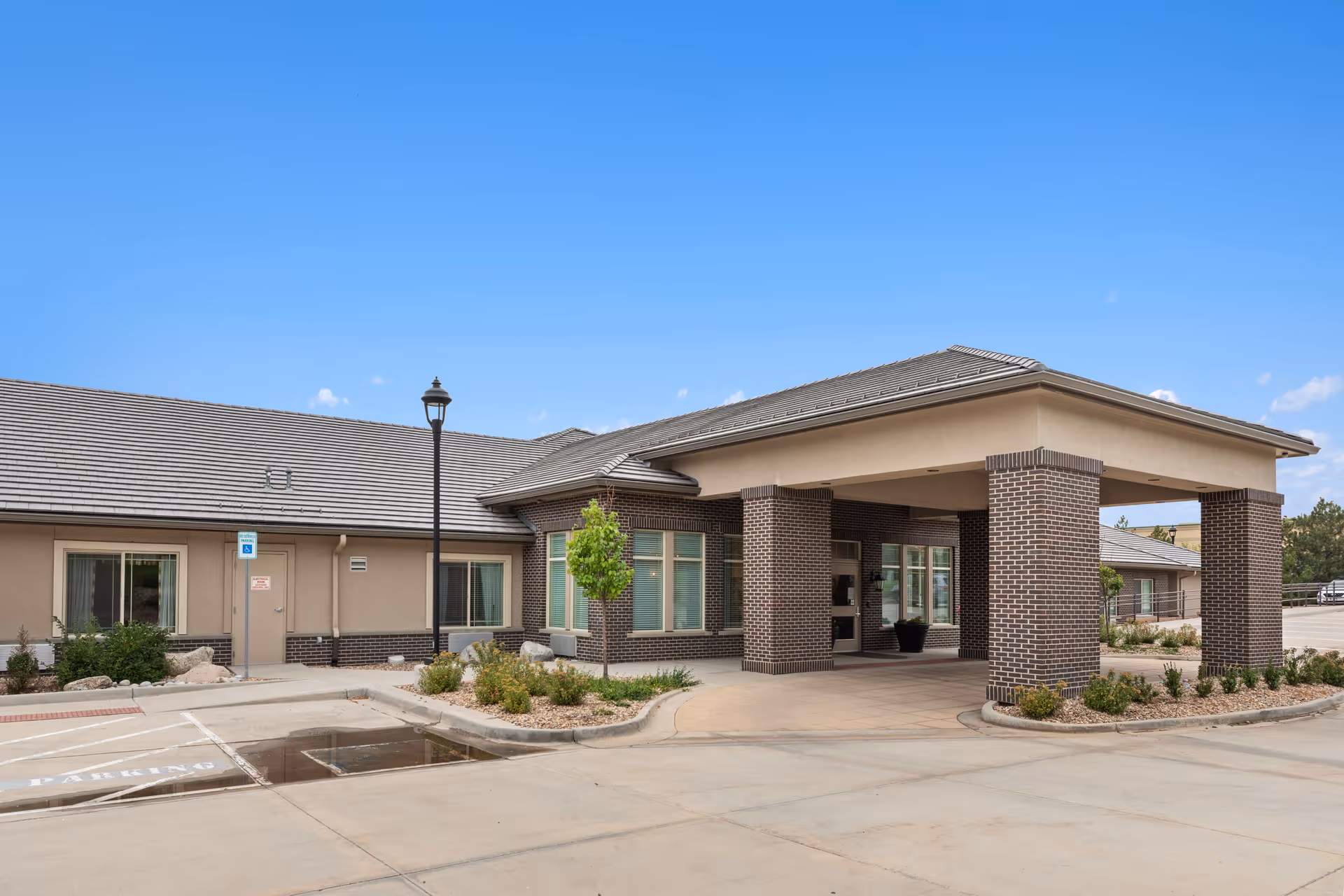 Exterior view of Belleview Heights Memory Care & Transitional Assisted Living facility showing the entrance with a covered drop-off area supported by brick columns, surrounded by landscaped plants and a clear blue sky.