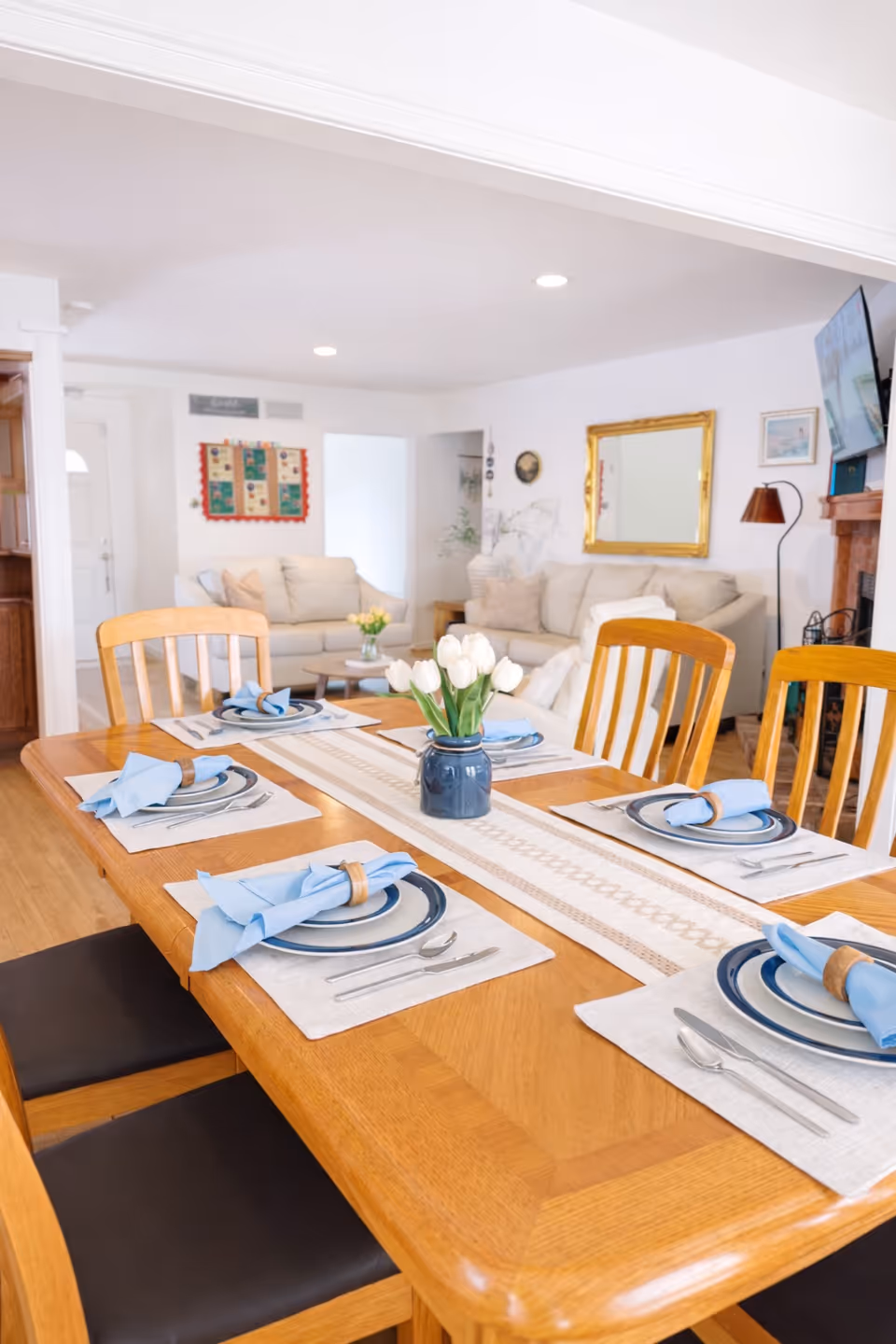 A wooden dining table with blue napkins and place settings in a bright living area.