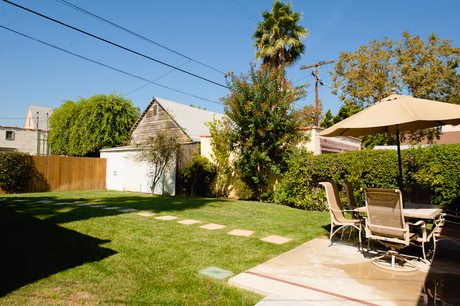A sunny backyard patio with chairs, umbrella, and a green lawn beside a shed.