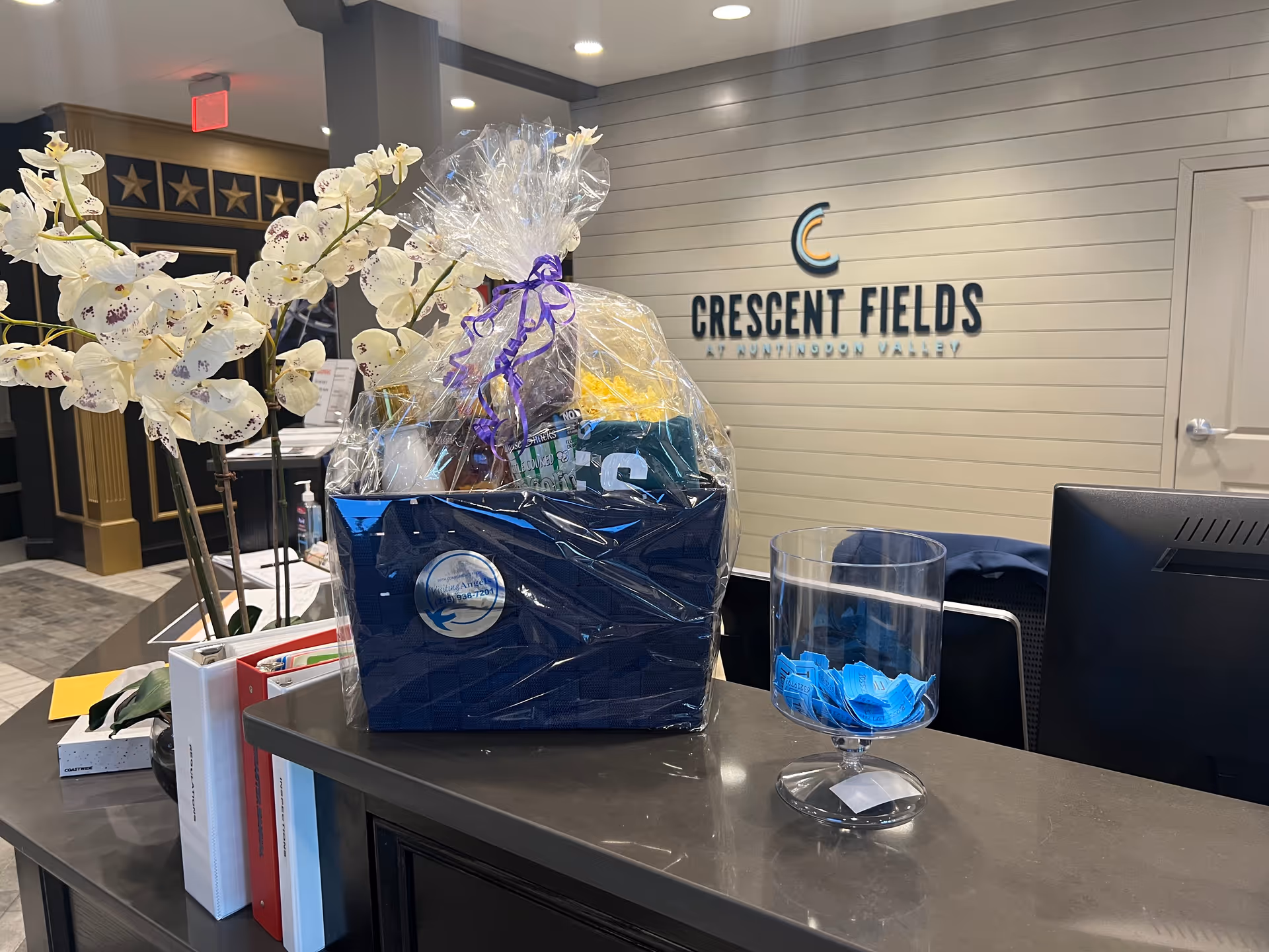 Reception desk with gift basket, orchid plant, and Crescent Fields sign on a gray paneled wall.