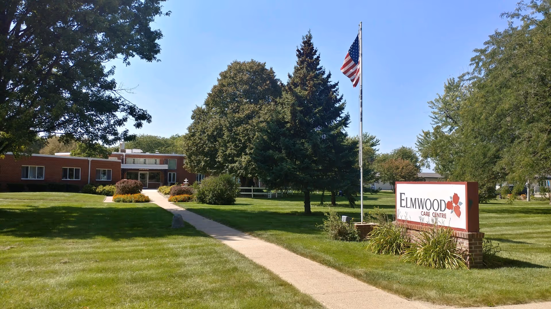 Well-kept facility exterior with brick building, manicured lawn, flagpole, and Elmwood Care Centre sign.