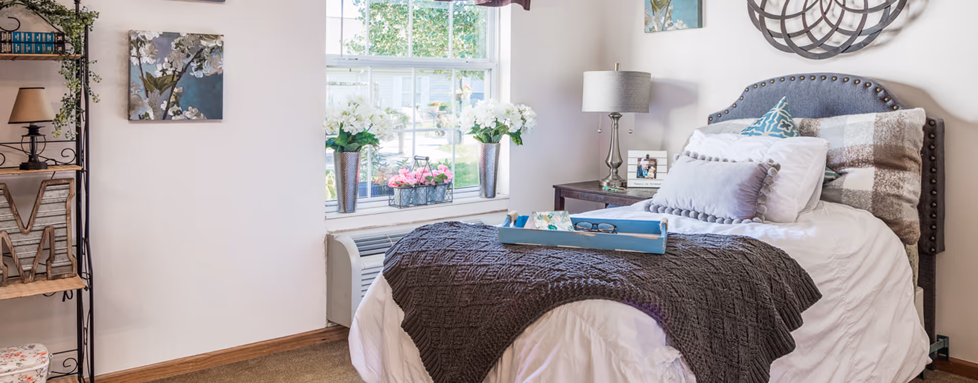 Cozy senior living bedroom with a bed, pillows, tray, and decorative shelf by a sunny window.