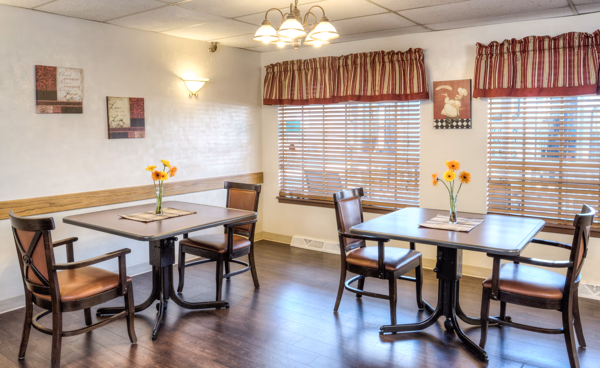 Two small square dining tables with wooden chairs and vases of yellow flowers in a well-lit room with window blinds and striped valances.