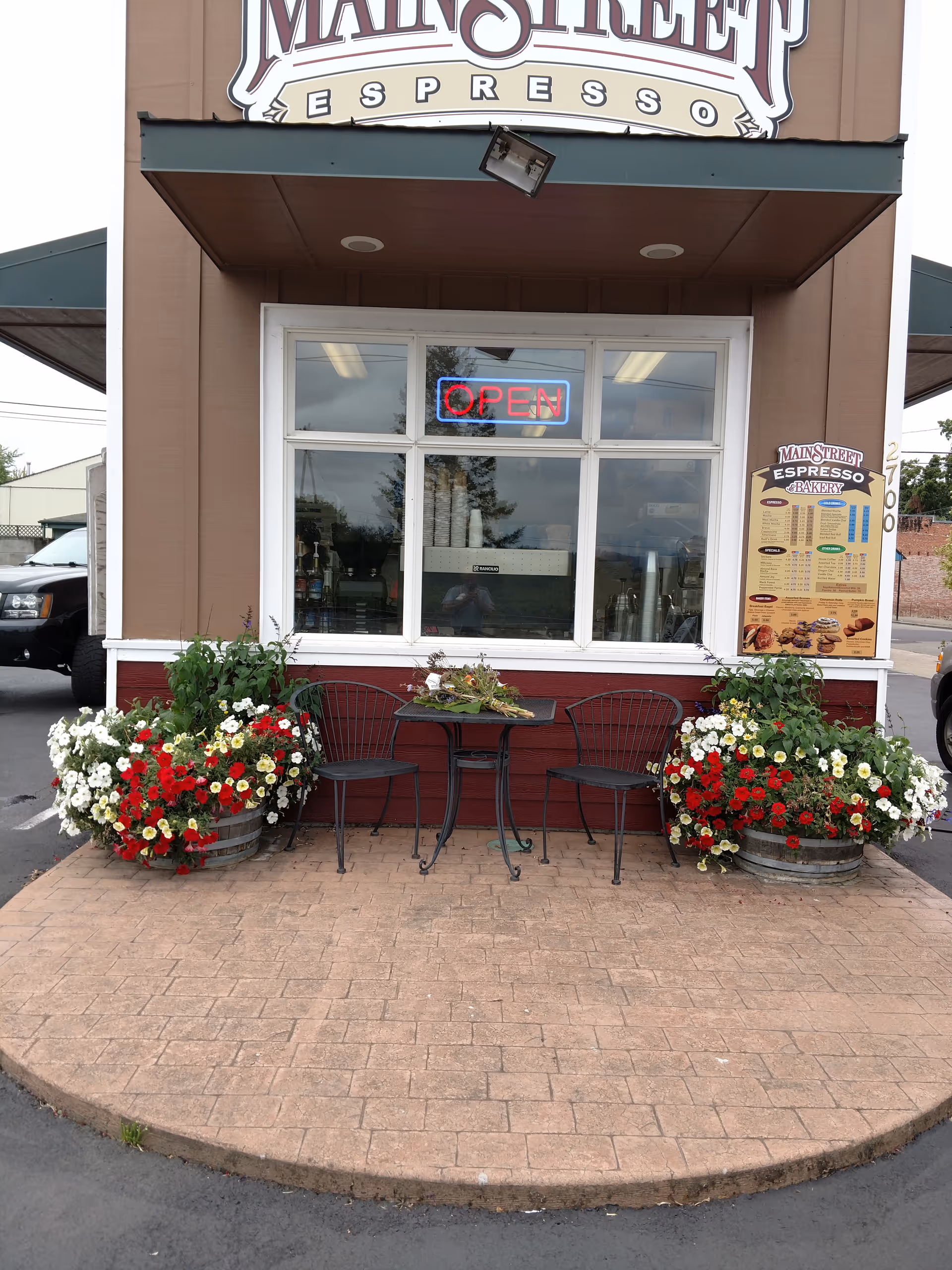 Front of a cafe with outdoor seating, flower pots, and an open neon sign.