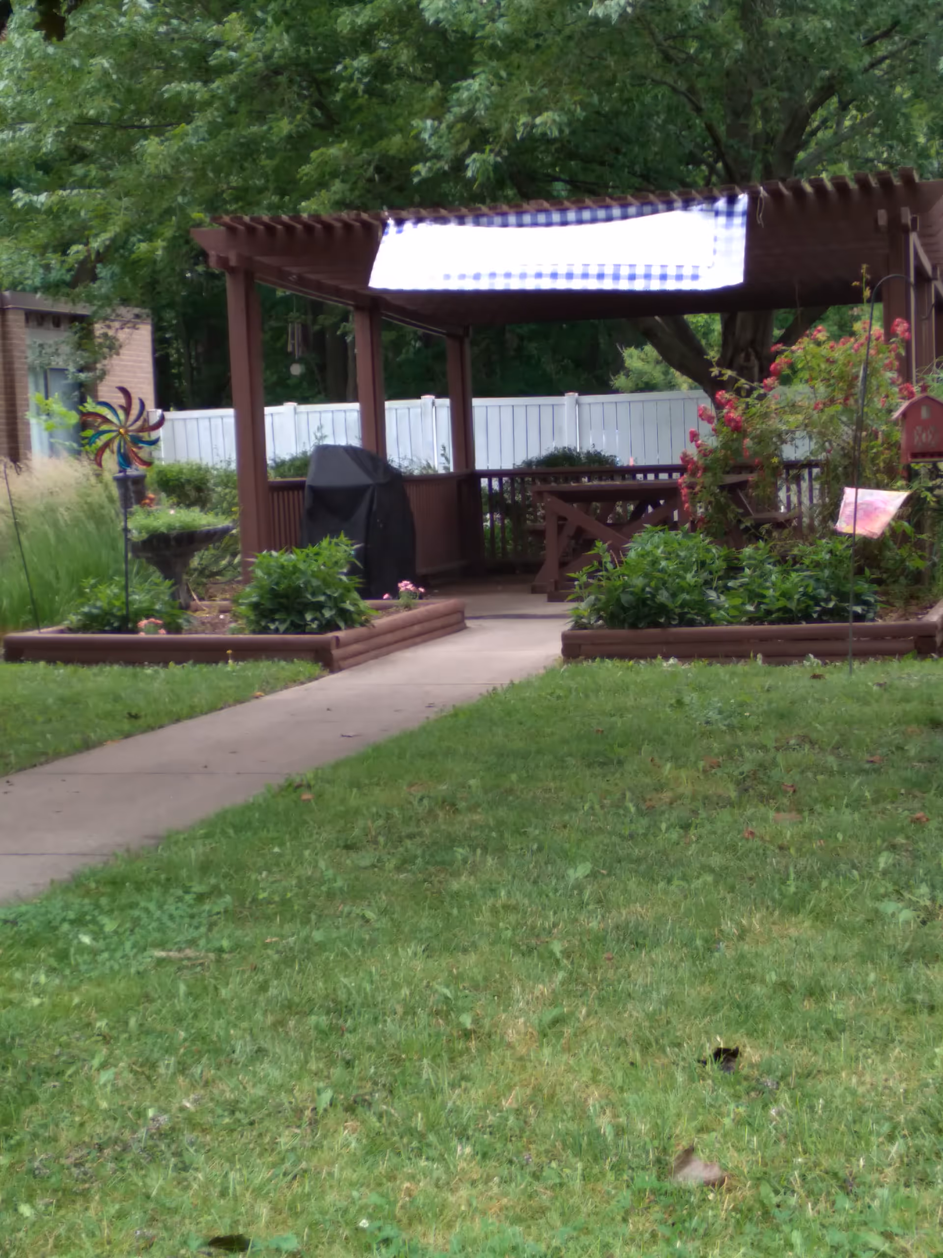 Outdoor backyard patio with wooden pergola, grill, garden beds, and lush green lawn.