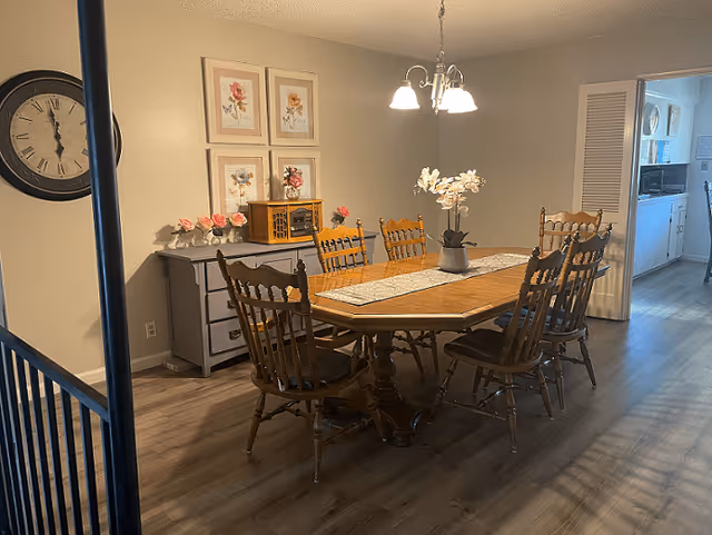 A dining room with a wooden dining table surrounded by six wooden chairs. A decorative runner and a potted white orchid are on the table. On the left wall, there is a large round clock and a sideboard with floral decorations and framed flower artwork above it. The room has wood flooring and neutral-colored walls. A doorway on the right leads to a kitchen area.