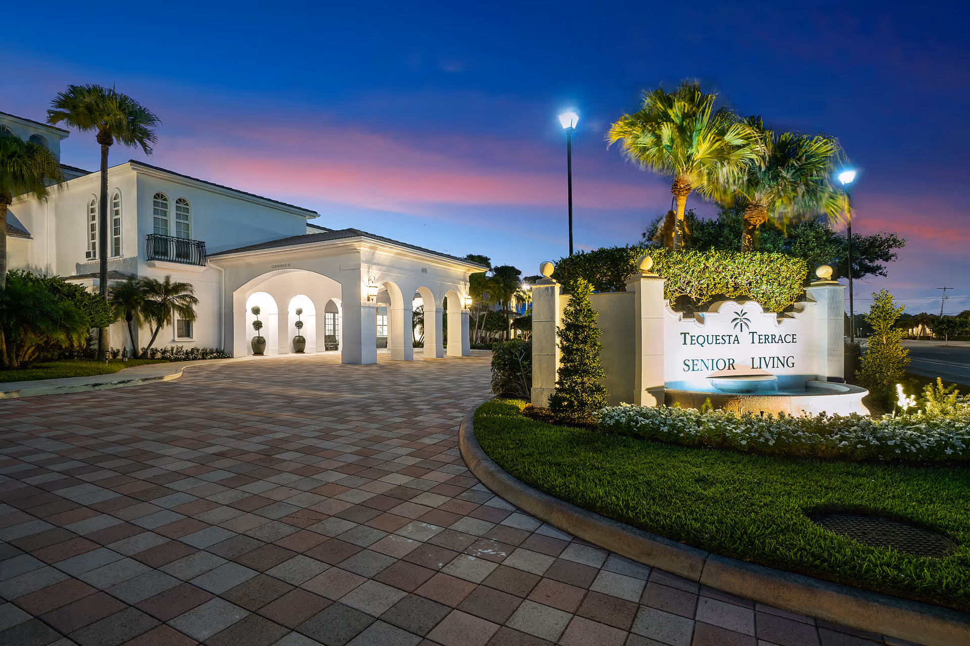 Exterior view of Tequesta Terrace Senior Living entrance with sign and arches at dusk.