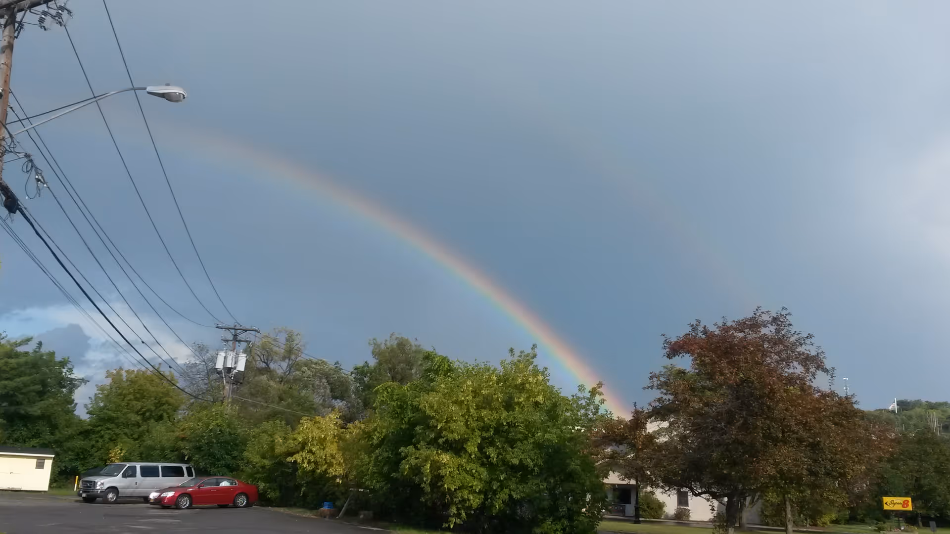 Double rainbow arcs over a parking lot beside a building with trees.